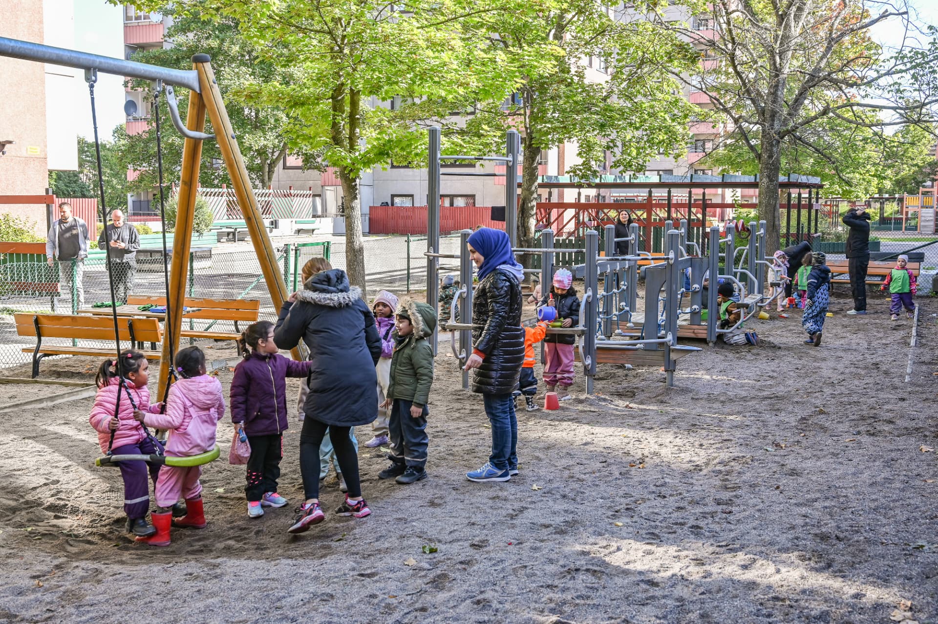 Barn och pedagoger på förskolans lekplats med gungor, klätterställning och stora träd